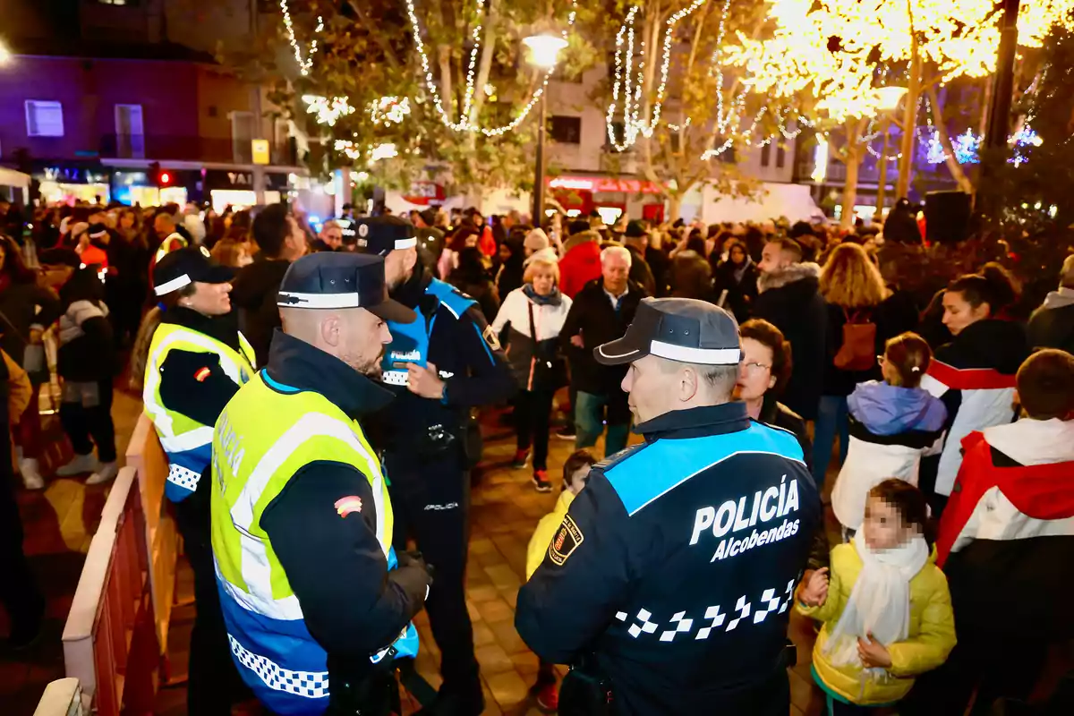 Un grupo de policías supervisa una multitud en un evento nocturno con luces decorativas en los árboles.