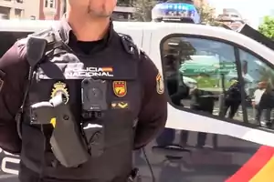 Un agente de la Policía Nacional de España con uniforme y equipo de servicio, de pie junto a un vehículo policial.