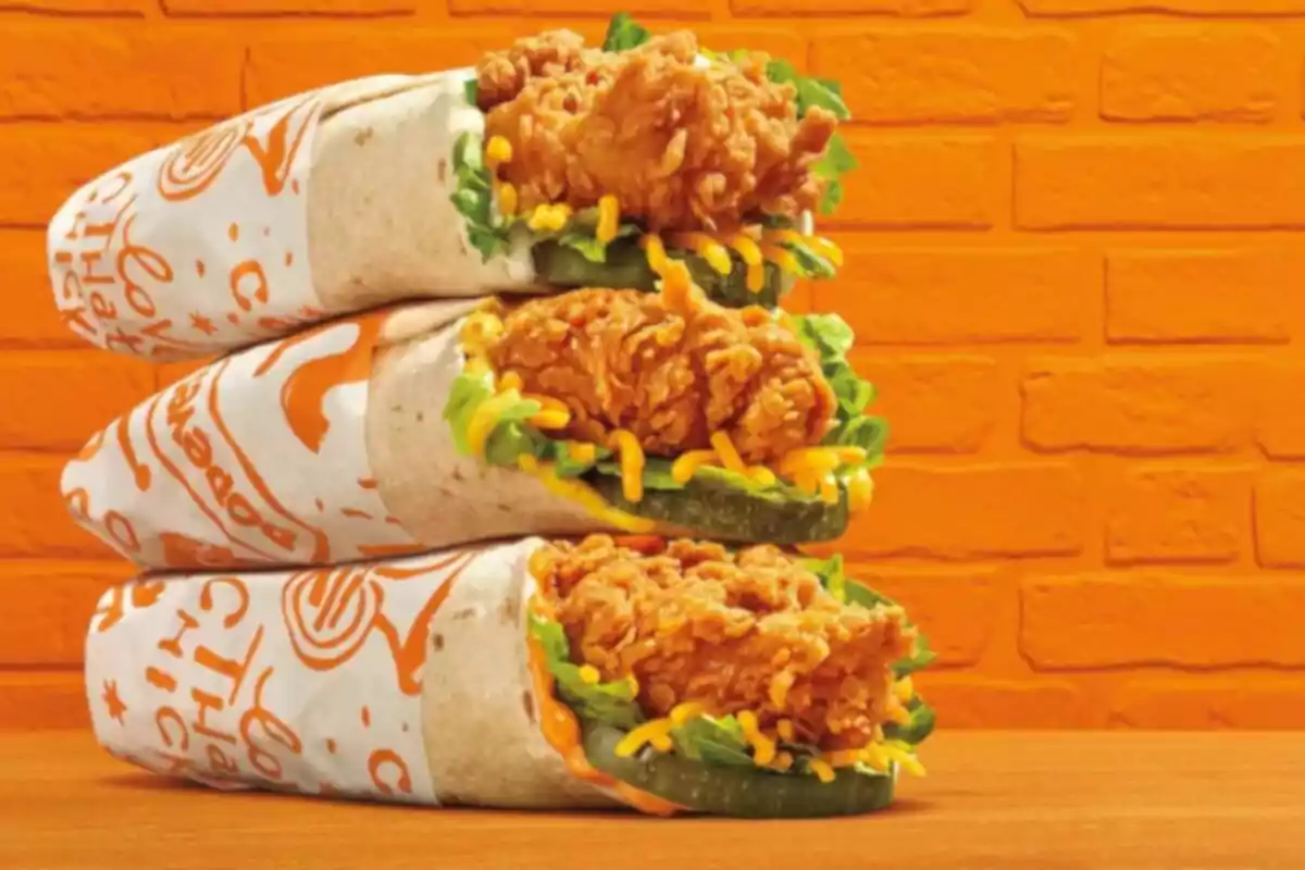 Tres burritos envueltos en papel con pollo frito, lechuga, pepinillos y queso rallado apilados sobre una mesa frente a una pared naranja Tres burritos envueltos en papel con pollo frito, lechuga, pepinillos y queso rallado apilados sobre una mesa frente a una pared naranja