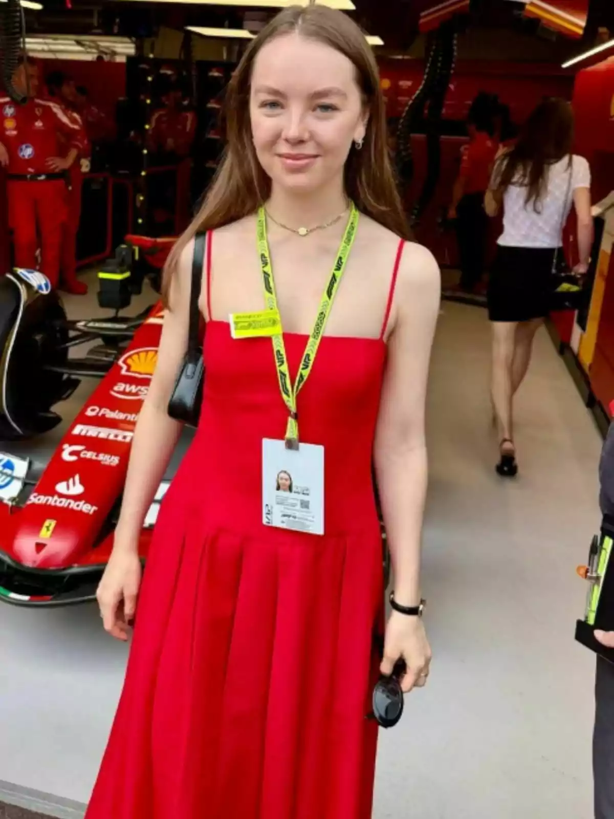 Alejandra of Hanover in a red dress with a badge hanging around her neck is standing in front of a race car in a garage. Alejandra of Hanover in a red dress with a badge hanging around her neck is standing in front of a race car in a garage.