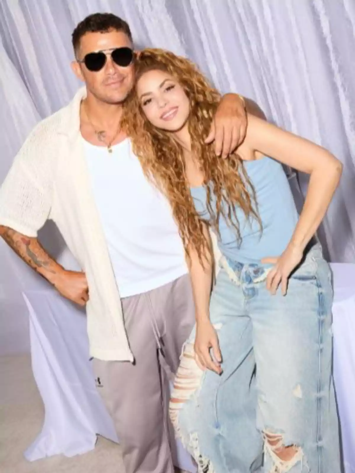 Alejandro Sanz and Shakira pose together, one with sunglasses and casual clothes, the other with curly hair and broken jeans, both smile in front of a light background. Alejandro Sanz and Shakira pose together, one with sunglasses and casual clothes, the other with curly hair and broken jeans, both smile in front of a light background.