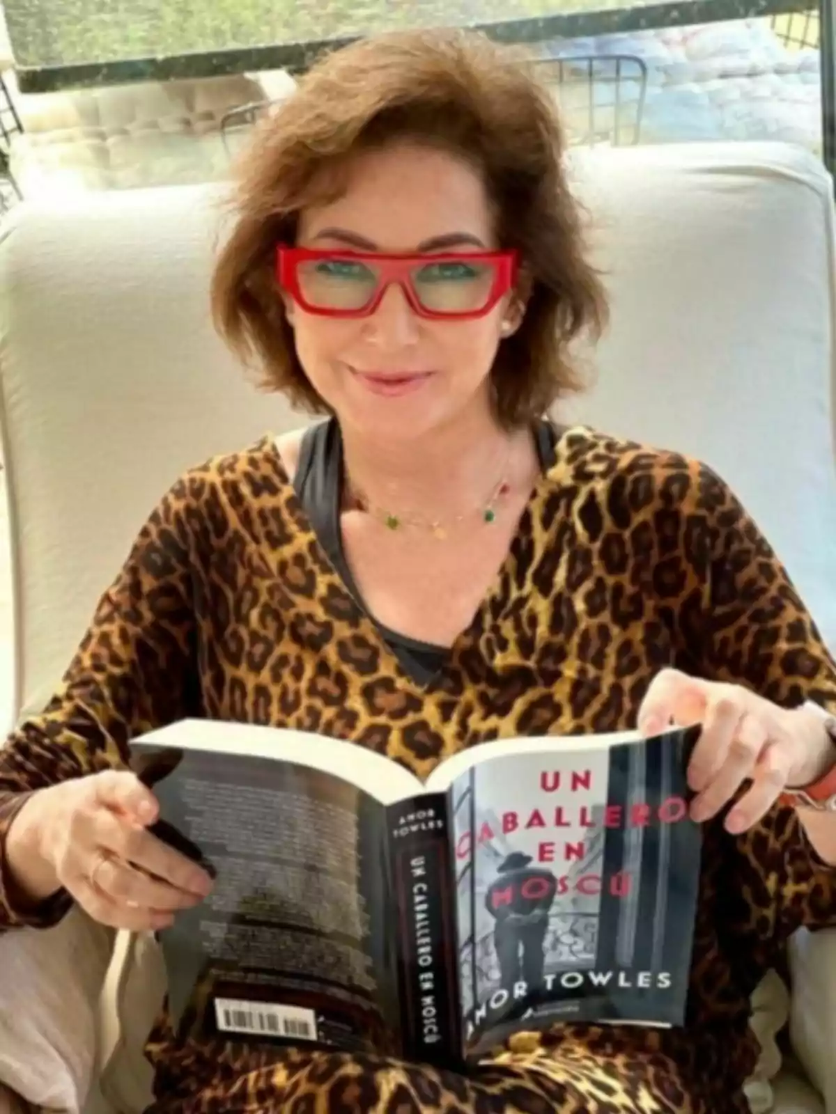 Ana Rosa Quintana con gafas rojas y un vestido de estampado animal está sentada leyendo un libro titulado Ana Rosa Quintana con gafas rojas y un vestido de estampado animal está sentada leyendo un libro titulado