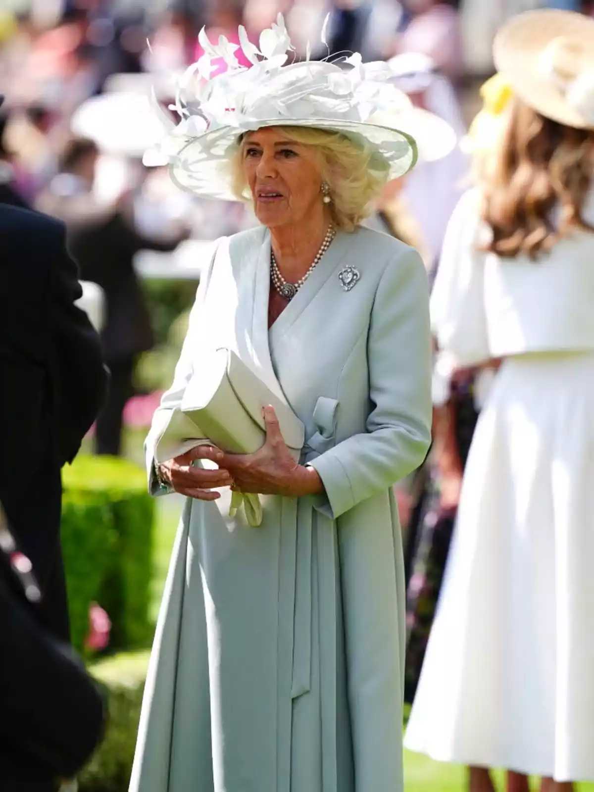Queen Camilla elegantly dressed in a light blue coat and a hat decorated with white flowers, holding a handbag at an outdoor event