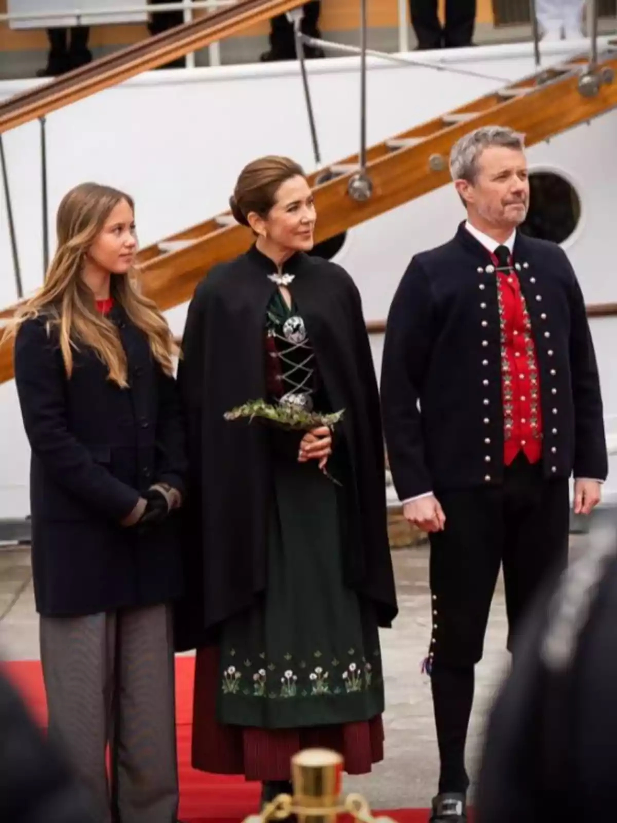 Josefina, Mary y Federico de Dinamarca de pie sobre una alfombra roja, vestidas con ropa formal y tradicional, posando frente a un barco Josefina, Mary y Federico de Dinamarca de pie sobre una alfombra roja, vestidas con ropa formal y tradicional, posando frente a un barco