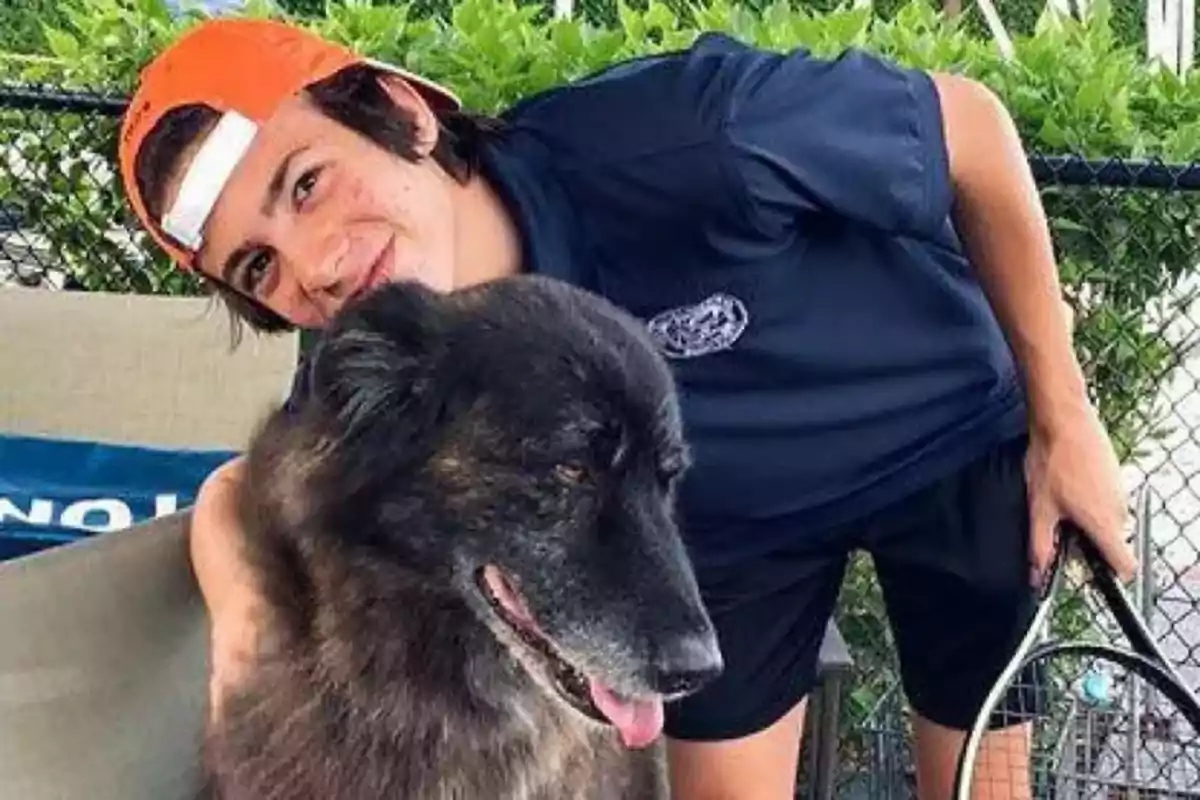 Guillermo Iglesias con gorra naranja al revés sonríe mientras abraza a un perro negro y peludo en un entorno al aire libre. Guillermo Iglesias con gorra naranja al revés sonríe mientras abraza a un perro negro y peludo en un entorno al aire libre.