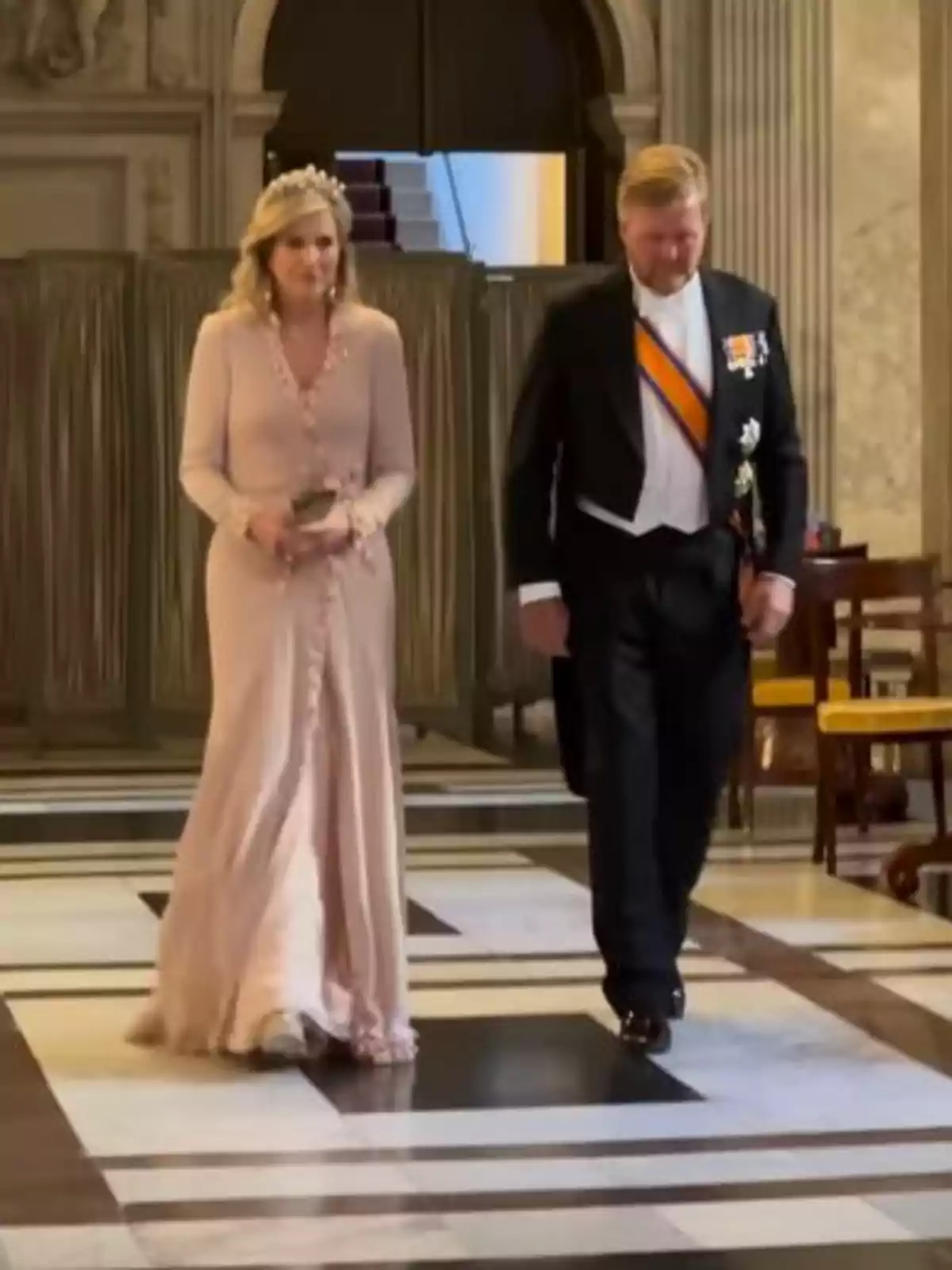 Máxima y Guillermo de Holanda elegantemente vestida camina por un salón con un suelo de mármol, ella lleva un vestido largo y una tiara, mientras que él viste un traje formal con condecoraciones. Máxima y Guillermo de Holanda elegantemente vestida camina por un salón con un suelo de mármol, ella lleva un vestido largo y una tiara, mientras que él viste un traje formal con condecoraciones.