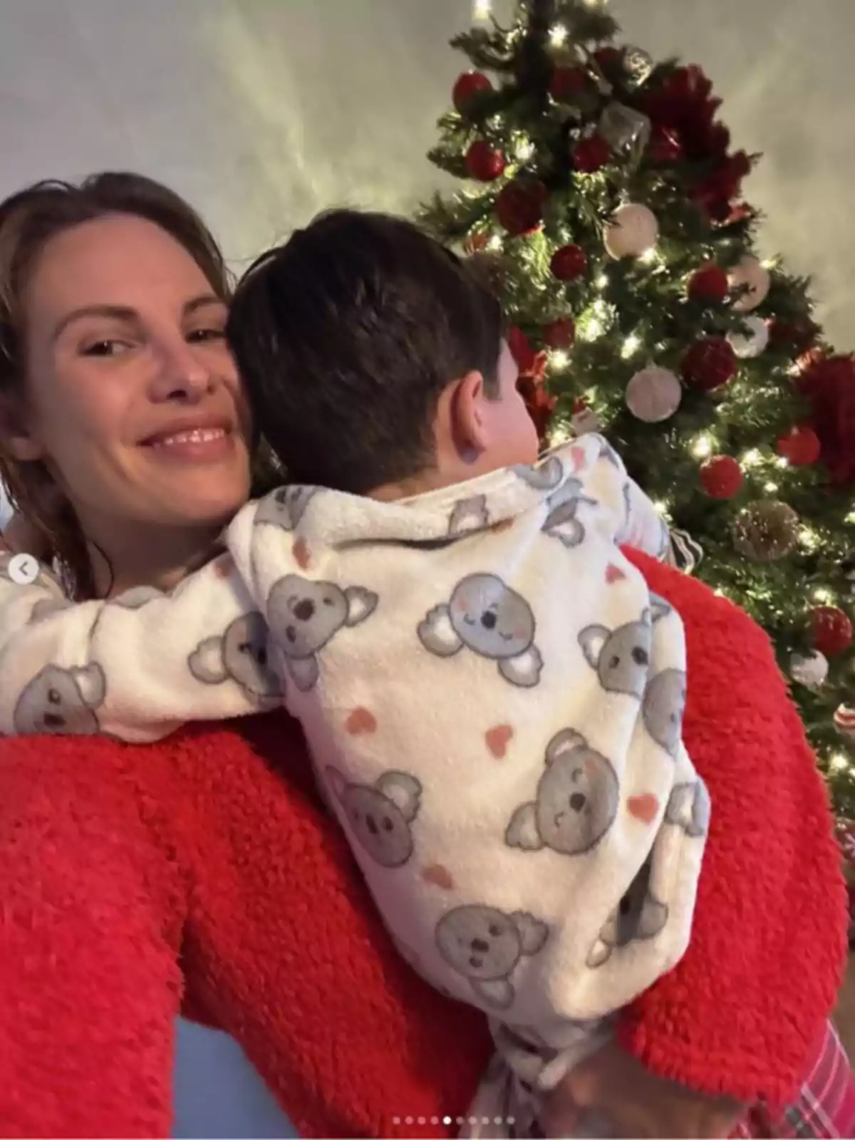 Jessica Bueno sonriente sostiene a un niño pequeño frente a un árbol de Navidad decorado con luces y adornos rojos. Jessica Bueno sonriente sostiene a un niño pequeño frente a un árbol de Navidad decorado con luces y adornos rojos.