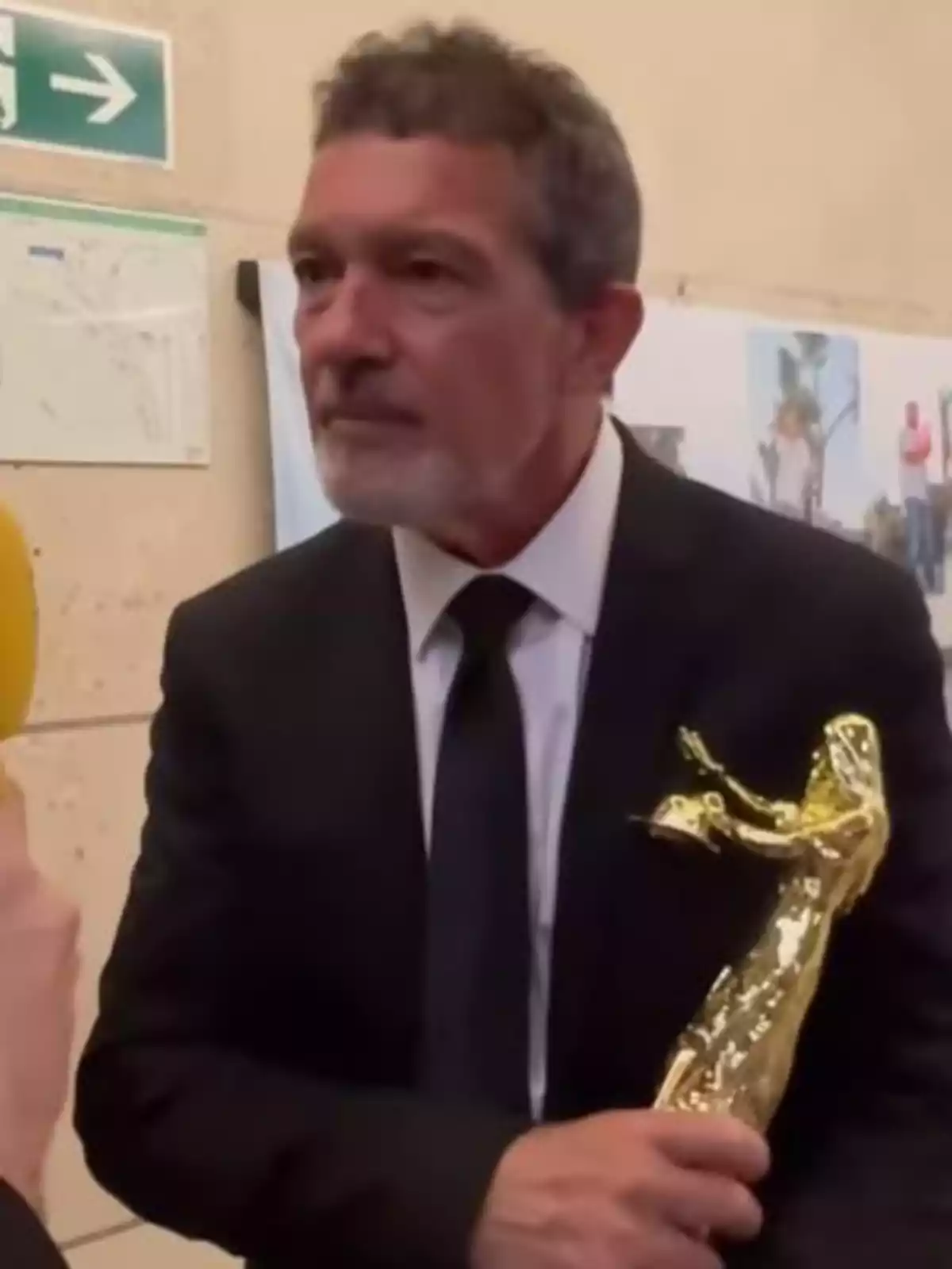 Antonio Banderas con traje oscuro sostiene un trofeo dorado mientras es entrevistado. Antonio Banderas con traje oscuro sostiene un trofeo dorado mientras es entrevistado.