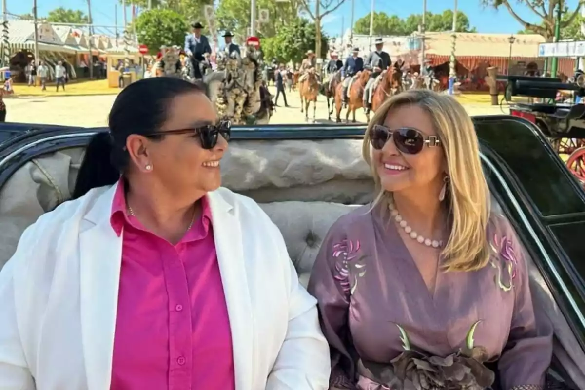 María del Monte e Inmaculada Casal sonrientes con gafas de sol están sentadas en un carruaje durante un evento al aire libre, con personas a caballo y decoraciones festivas al fondo. María del Monte e Inmaculada Casal sonrientes con gafas de sol están sentadas en un carruaje durante un evento al aire libre, con personas a caballo y decoraciones festivas al fondo.
