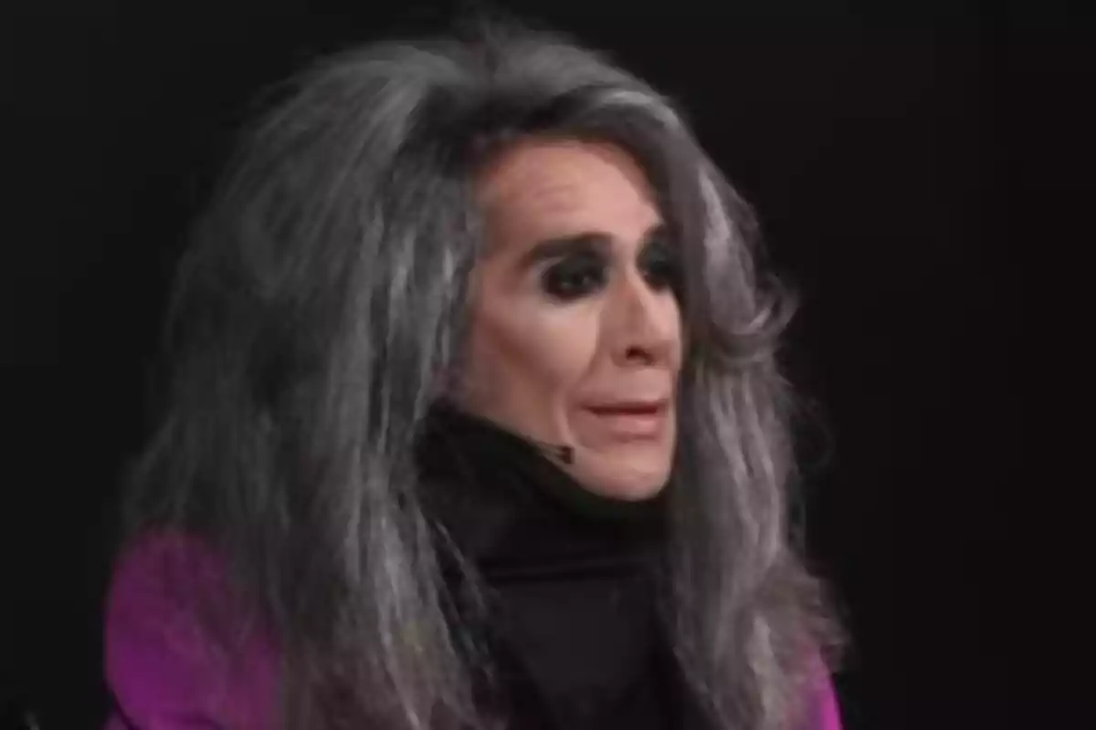 Mario Vaquerizo con cabello largo y canoso, maquillaje oscuro y vestimenta negra y morada, en un fondo oscuro. Mario Vaquerizo con cabello largo y canoso, maquillaje oscuro y vestimenta negra y morada, en un fondo oscuro.