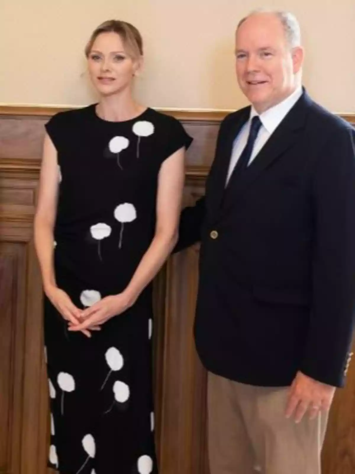 Charlene con vestido negro de lunares blancos y Alberto de Mónaco con saco oscuro y pantalón claro posan juntos en un interior elegante. Charlene con vestido negro de lunares blancos y Alberto de Mónaco con saco oscuro y pantalón claro posan juntos en un interior elegante.