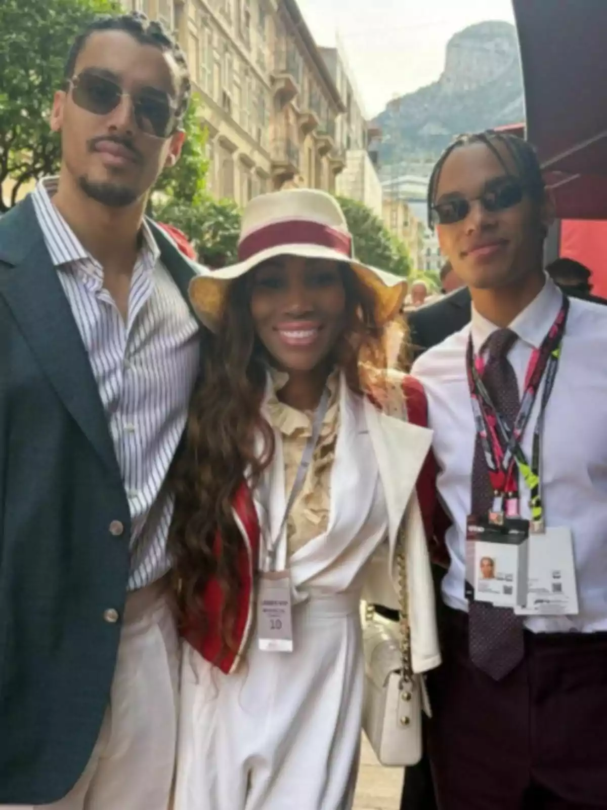 Tres personas posan sonrientes al aire libre en una calle elegante, una mujer en el centro lleva un sombrero de ala ancha y los dos hombres a su lado usan gafas de sol y ropa formal, al fondo se ven edificios y una montaña Tres personas posan sonrientes al aire libre en una calle elegante, una mujer en el centro lleva un sombrero de ala ancha y los dos hombres a su lado usan gafas de sol y ropa formal, al fondo se ven edificios y una montaña