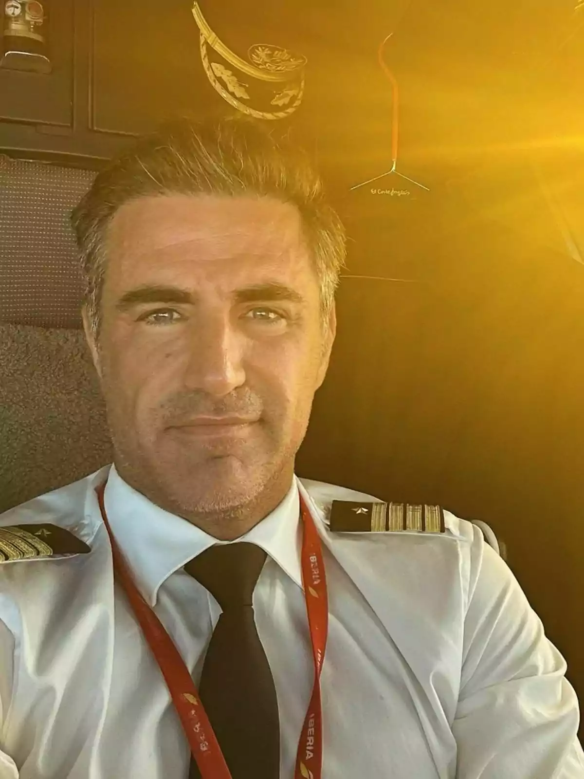 Perico Durán con uniforme y corbata negra está sentado en la cabina de un avión con luz dorada iluminando el espacio. Perico Durán con uniforme y corbata negra está sentado en la cabina de un avión con luz dorada iluminando el espacio.