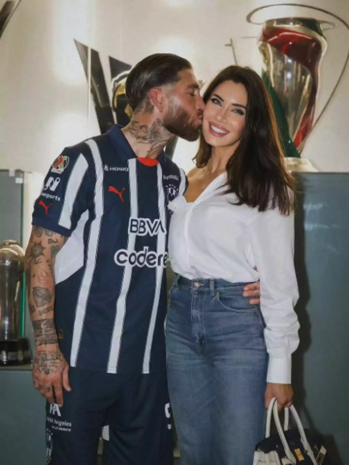 Sergio Ramos con uniforme de fútbol besa en la mejilla a Pilar Rubio que sonríe, ambos posan frente a trofeos. Sergio Ramos con uniforme de fútbol besa en la mejilla a Pilar Rubio que sonríe, ambos posan frente a trofeos.