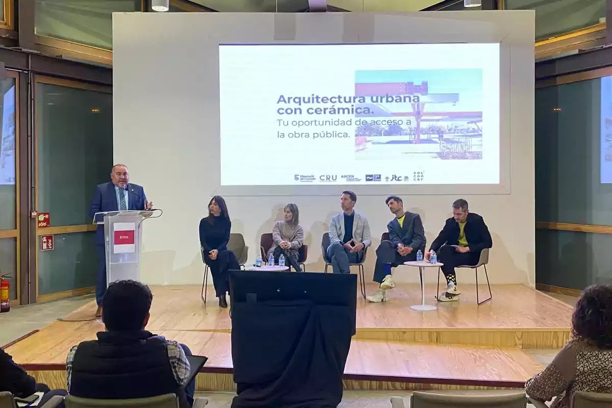 A group of people is participating in a conference on urban architecture with ceramics, featuring a speaker at a podium and a panel discussion on a stage.