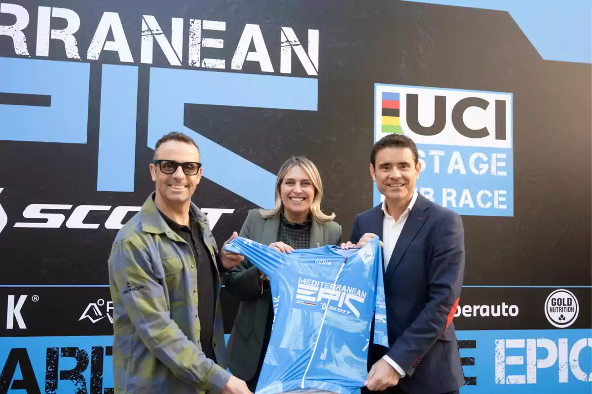 Three people are holding a blue jersey in front of a Mediterranean Epic UCI Stage Race banner.