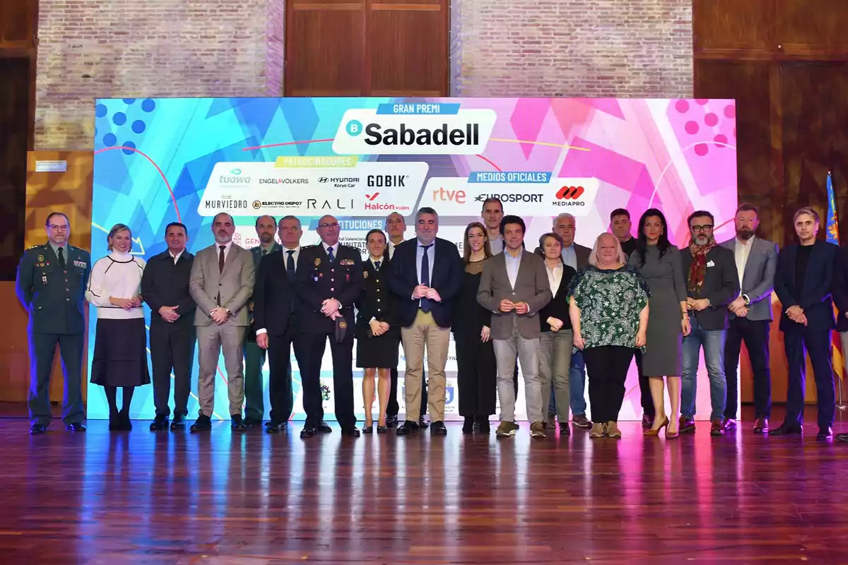 A group of people posing in front of a colorful sign with sponsor logos and official media.