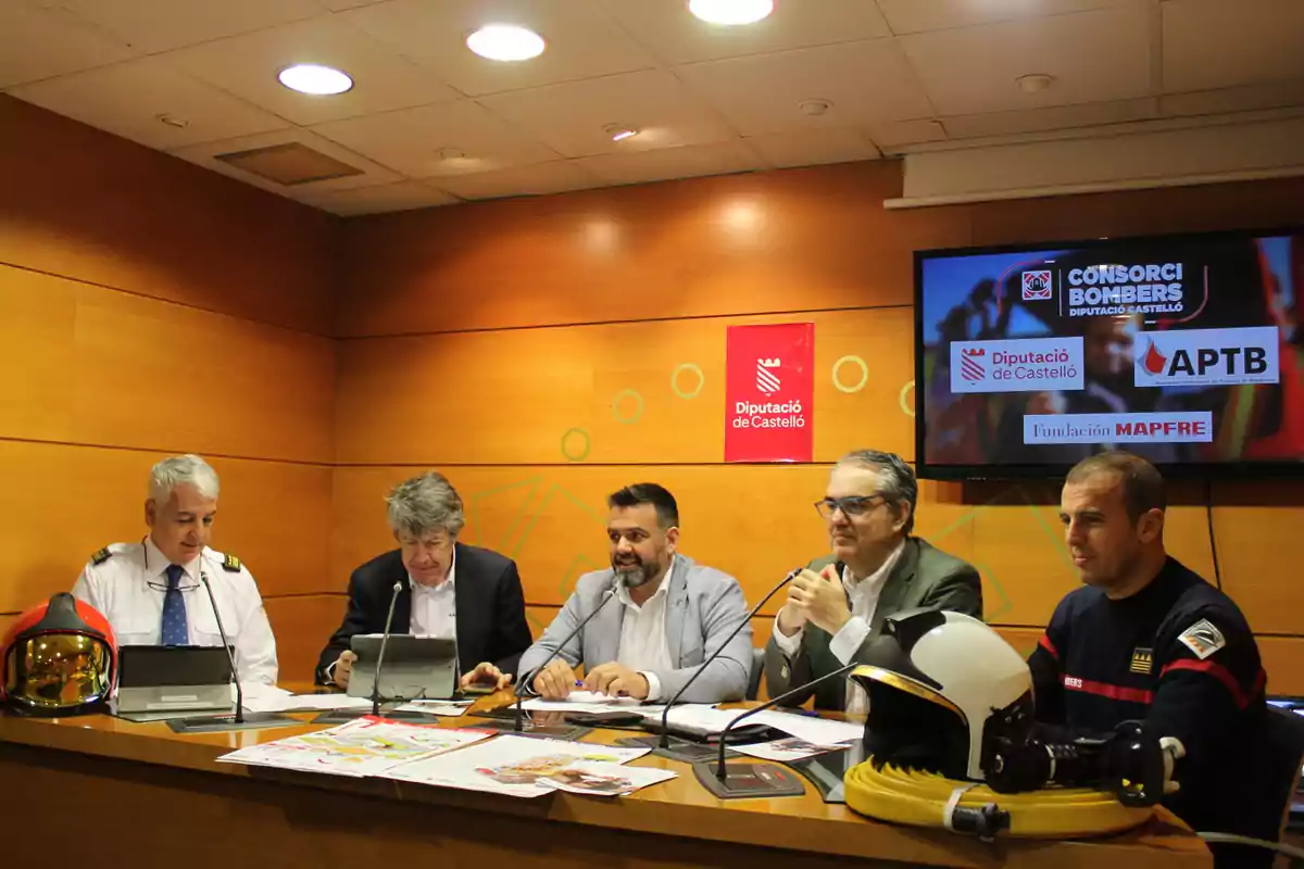 Five men seated at a table during a press conference in a room with wooden walls, wearing firefighter helmets and a monitor displaying logos of firefighter-related organizations.