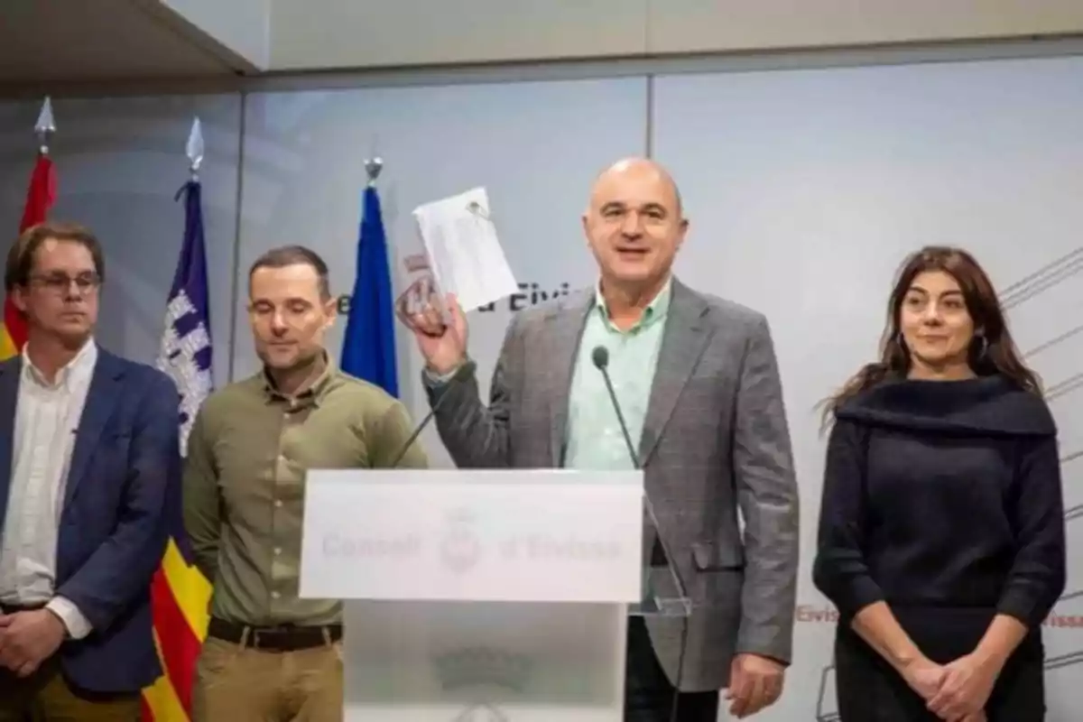 Un grupo de personas en una conferencia de prensa con banderas de fondo y un hombre sosteniendo documentos.