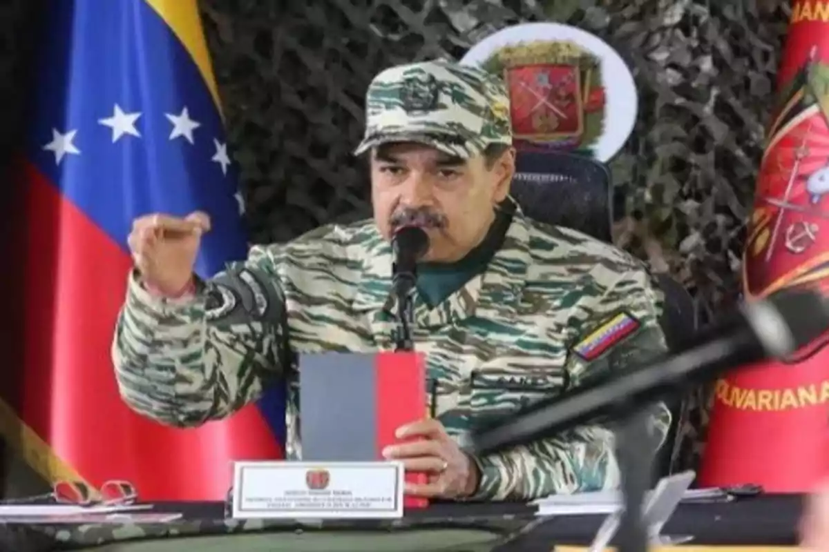 Un hombre con uniforme militar camuflado habla en un micrófono mientras sostiene un libro, con la bandera de Venezuela y un escudo en el fondo.