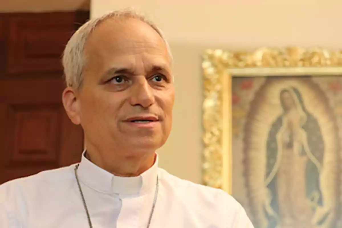 Un hombre con vestimenta clerical frente a una imagen religiosa enmarcada.