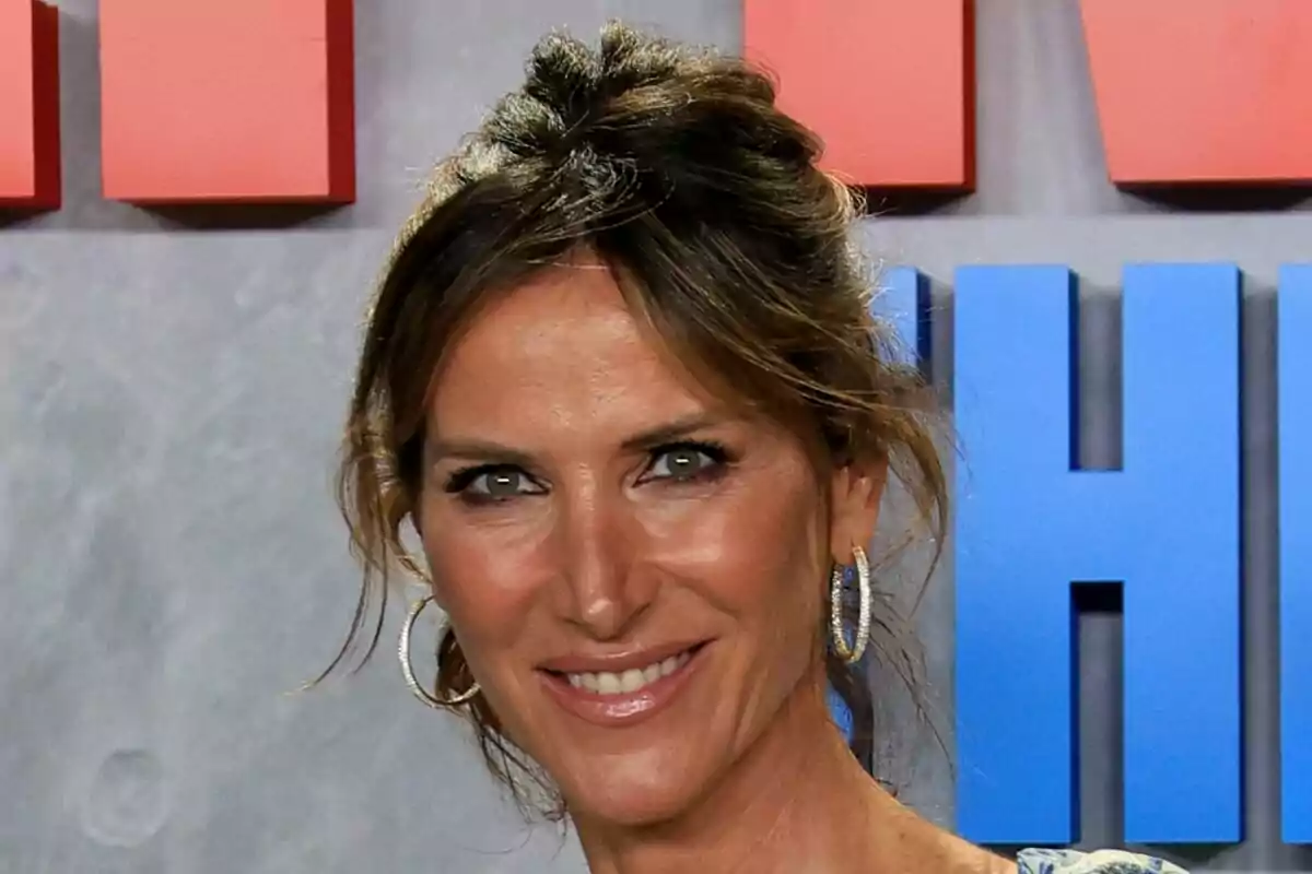 Alejandra Prat sonriendo con aretes grandes y cabello recogido frente a un fondo con letras rojas y azules.