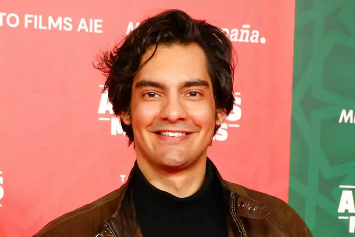 Alejandro Reyes sonriendo con una chaqueta marrón frente a un fondo rojo y verde con texto. Alejandro Reyes sonriendo con una chaqueta marrón frente a un fondo rojo y verde con texto.