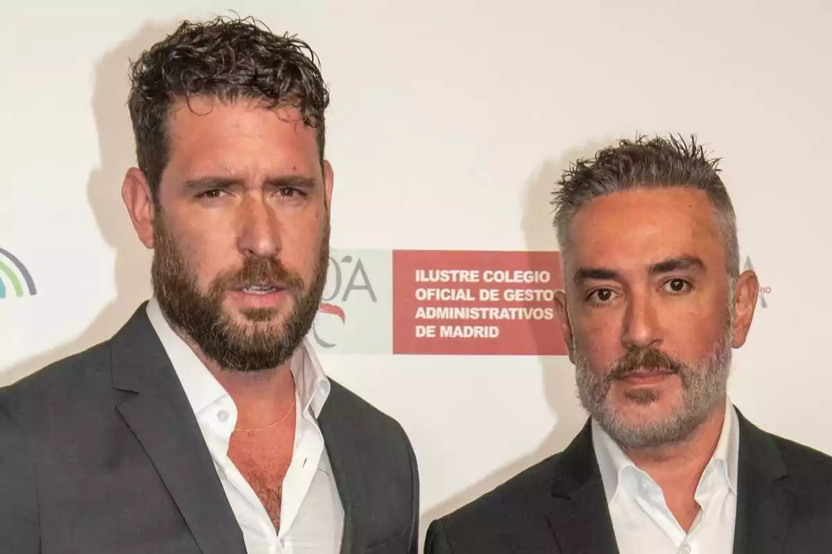 Fran Antón y Kiko Hernández con barba y traje posan frente a un fondo con logotipos y texto del Ilustre Colegio Oficial de Gestores Administrativos de Madrid.