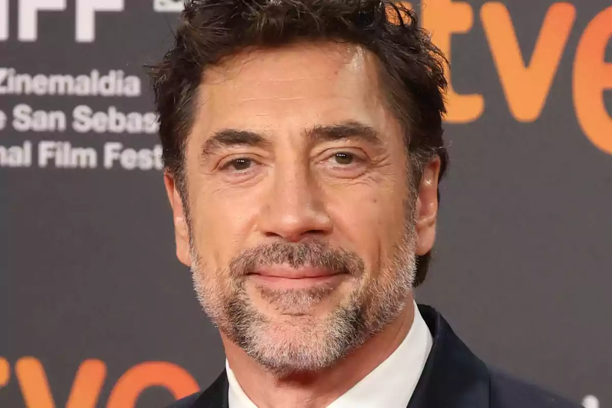 Javier Bardem con traje en un evento con fondo de letras y logotipos.