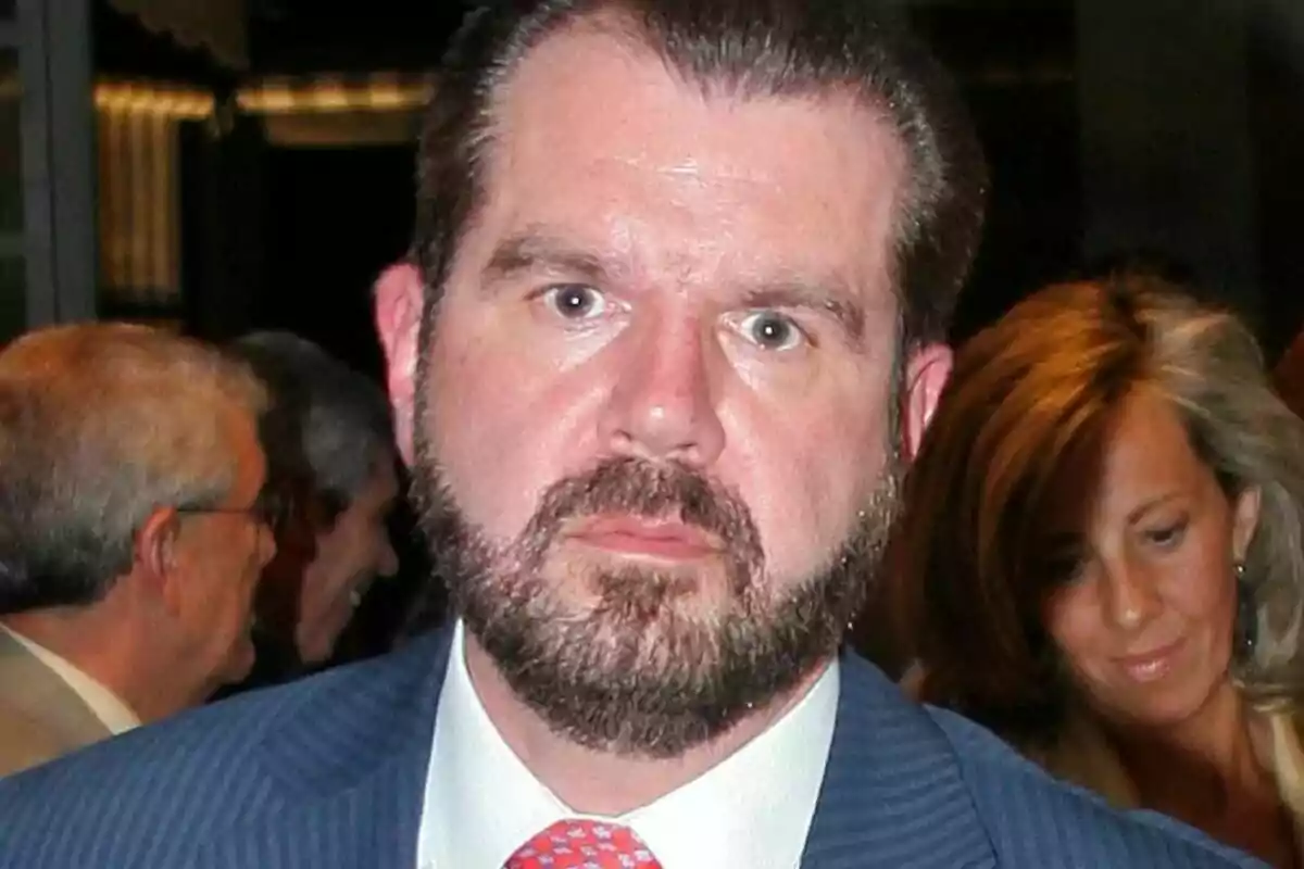 Jesus Ortiz with a beard and suit at a social event with people in the background.