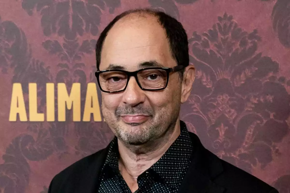 Jordi Sánchez con gafas y barba ligera, vestido con una camisa oscura y chaqueta, frente a un fondo decorativo.