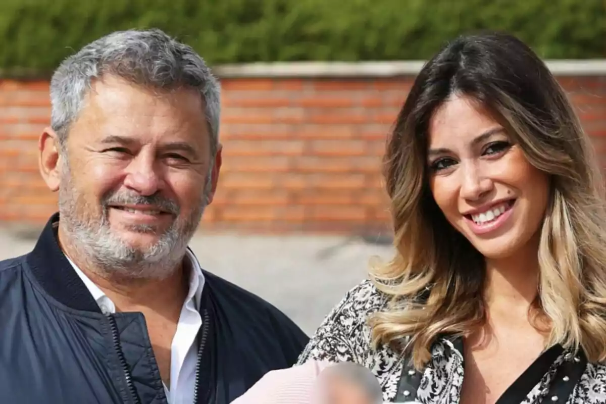 Miki Nadal y Helena Aldea sonríen a la cámara mientras sostienen a un bebé, con un fondo de ladrillos y vegetación.