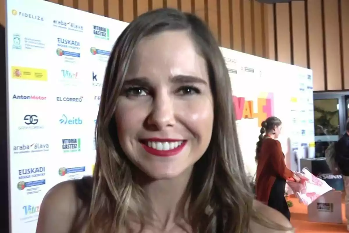 Natalia Sánchez sonriente en un evento con un fondo de logotipos y una persona en segundo plano. Natalia Sánchez sonriente en un evento con un fondo de logotipos y una persona en segundo plano.