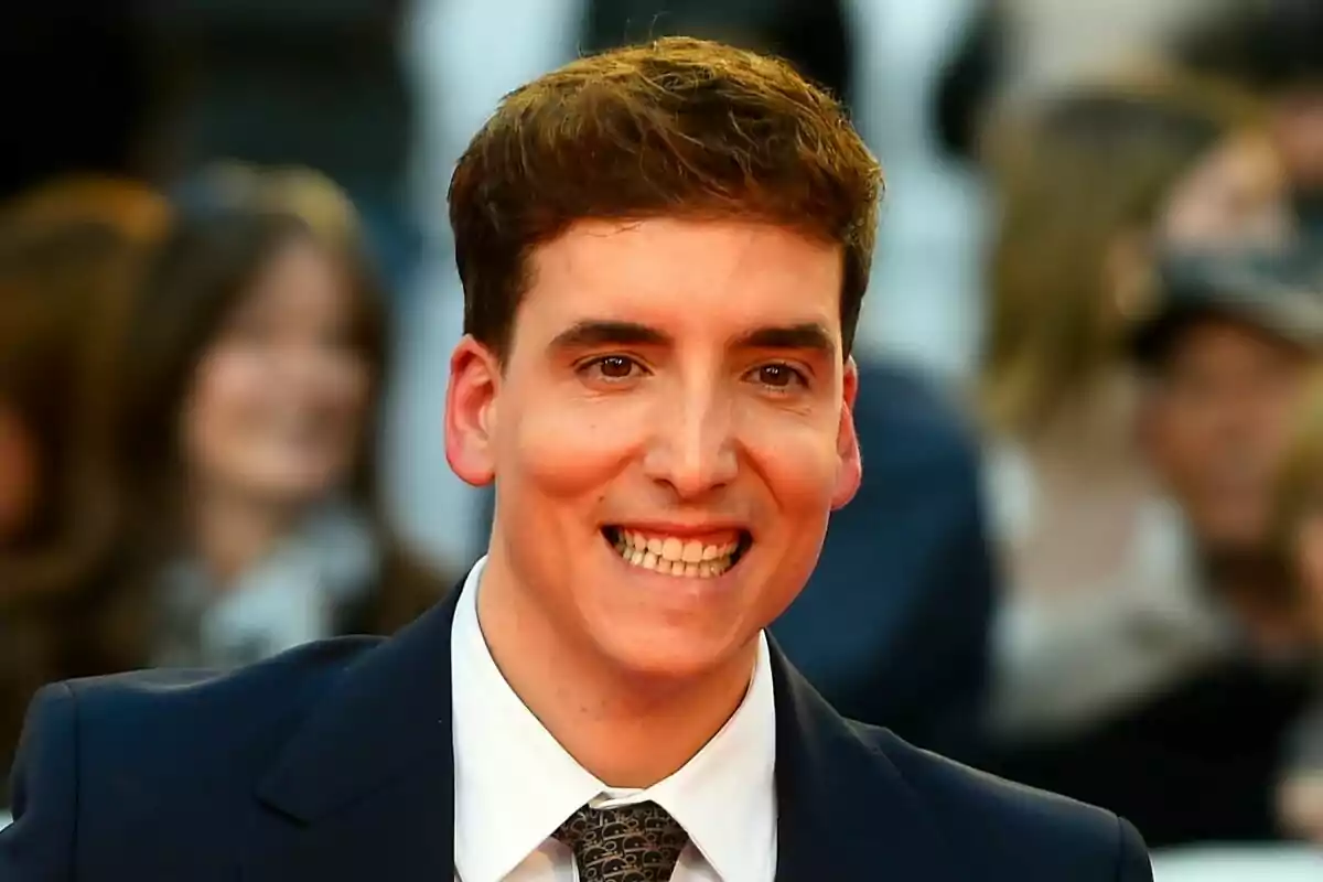 Xuso Jones con cabello castaño y sonrisa amplia, vistiendo traje oscuro y corbata, en un evento con fondo desenfocado de personas.