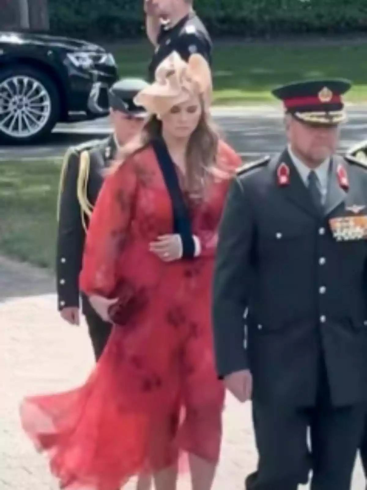 Princess Amalia of the Netherlands, dressed in a red dress and an elegant hat, walks alongside men in military uniforms at an outdoor event.