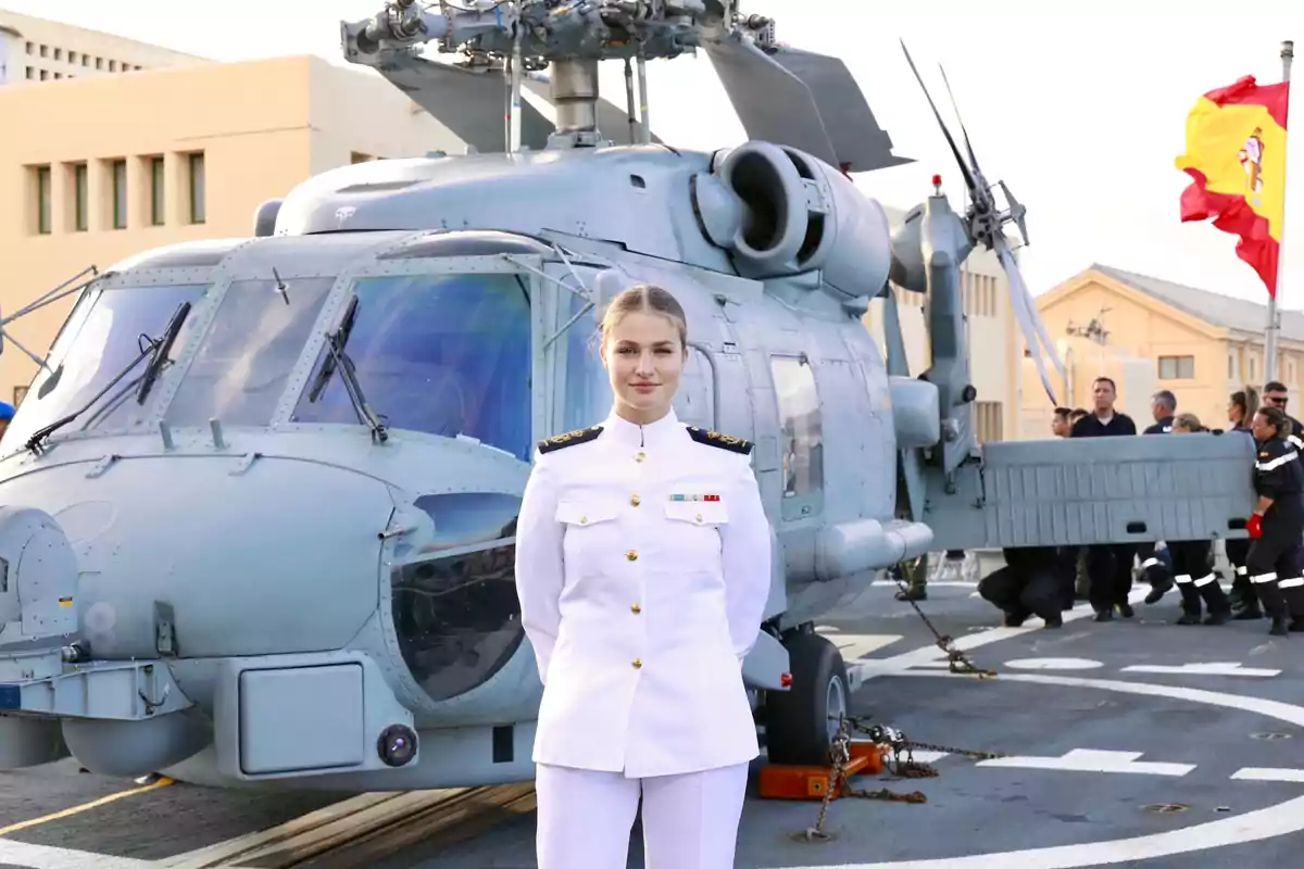 Leonor con uniforme militar blanco de gala posando frente a un helicóptero en una base naval con bandera de España al fondo Leonor con uniforme militar blanco de gala posando frente a un helicóptero en una base naval con bandera de España al fondo