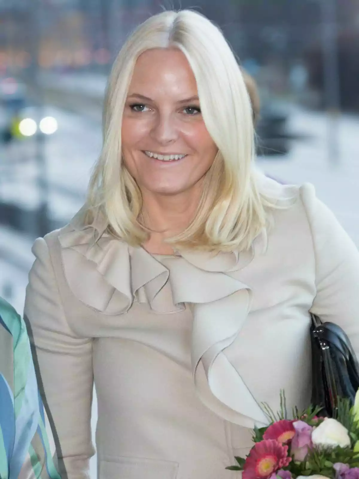 Princess Mette-Marit, wearing a light-coloured coat with ruffled details, holds a bouquet of flowers as she smiles.