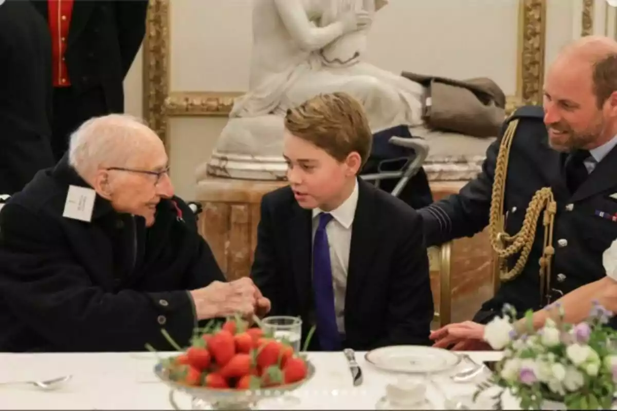 Three elegantly dressed people are sitting at a table decorated with strawberries and flowers, conversing in a formal setting.