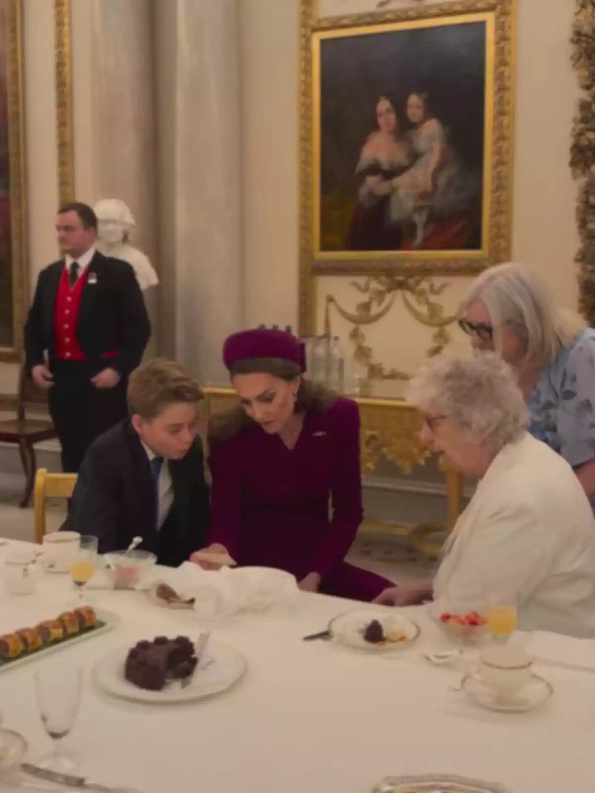 Several elegantly dressed people are seated around a table with desserts and drinks in a room decorated with paintings and columns.