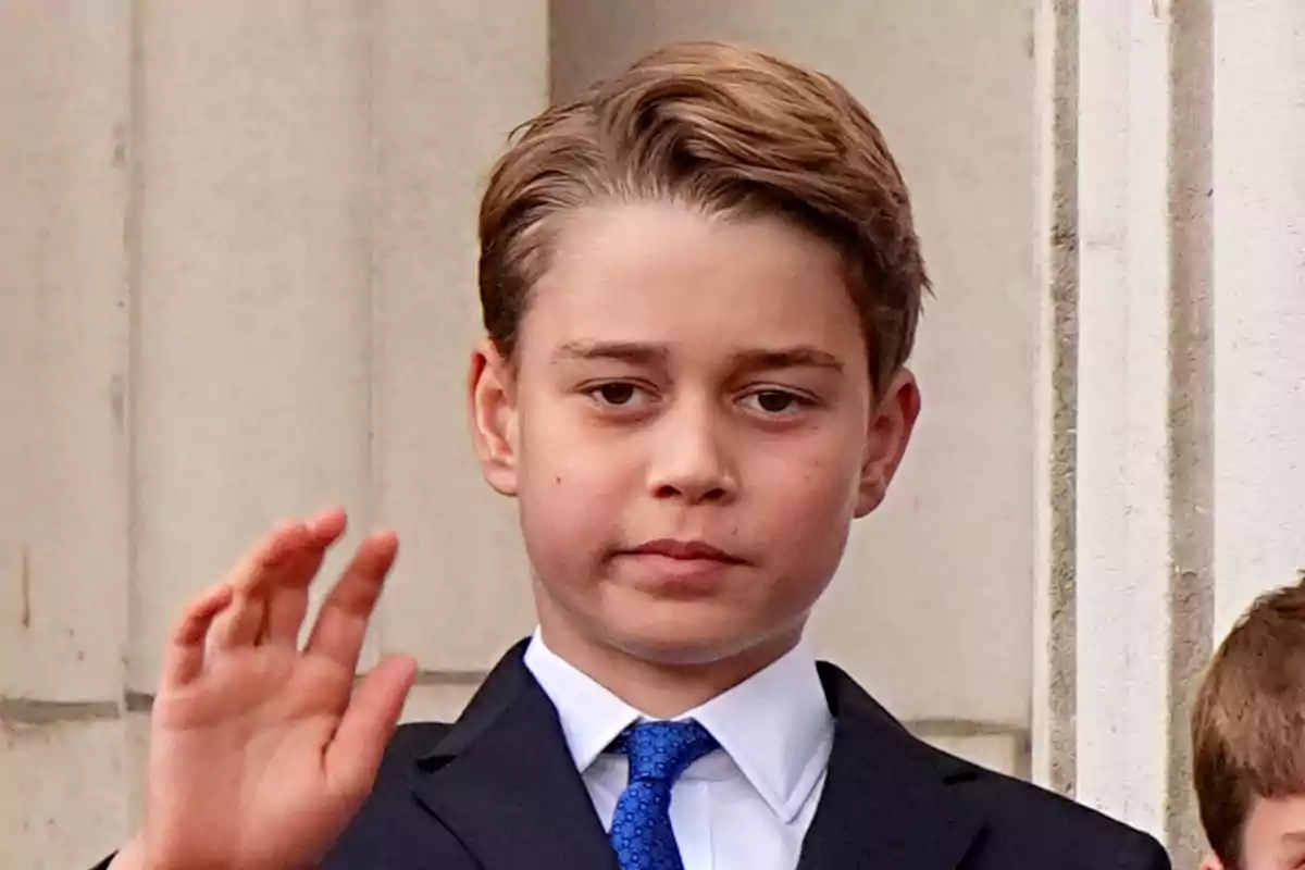 Prince George in a dark suit and blue tie raises his hand in front of a stone building. Prince George in a dark suit and blue tie raises his hand in front of a stone building.