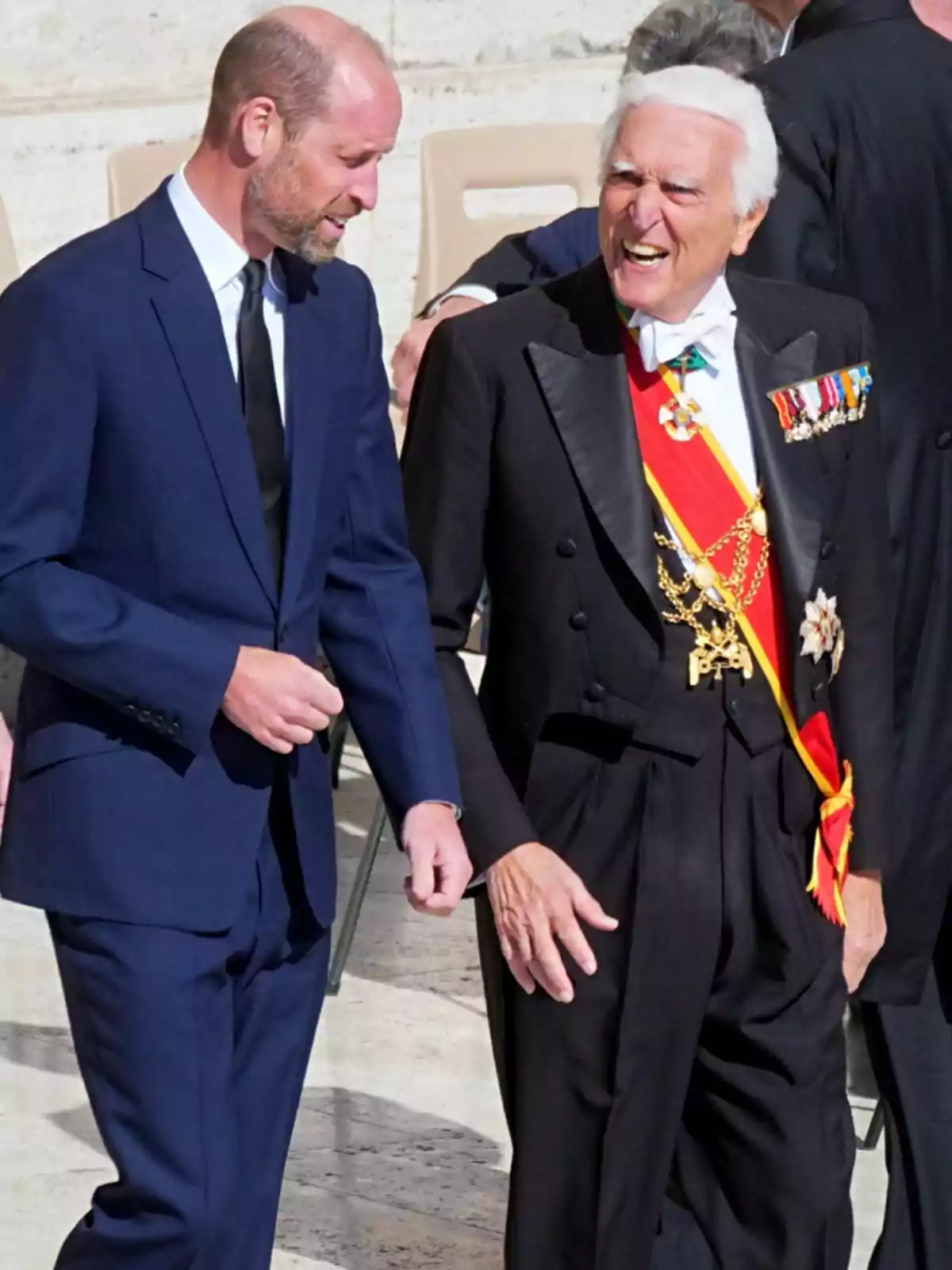 El príncipe Guillermo y otro hombre conversan animadamente; uno lleva un traje azul y el otro un traje formal con condecoraciones y una banda roja. El príncipe Guillermo y otro hombre conversan animadamente; uno lleva un traje azul y el otro un traje formal con condecoraciones y una banda roja.