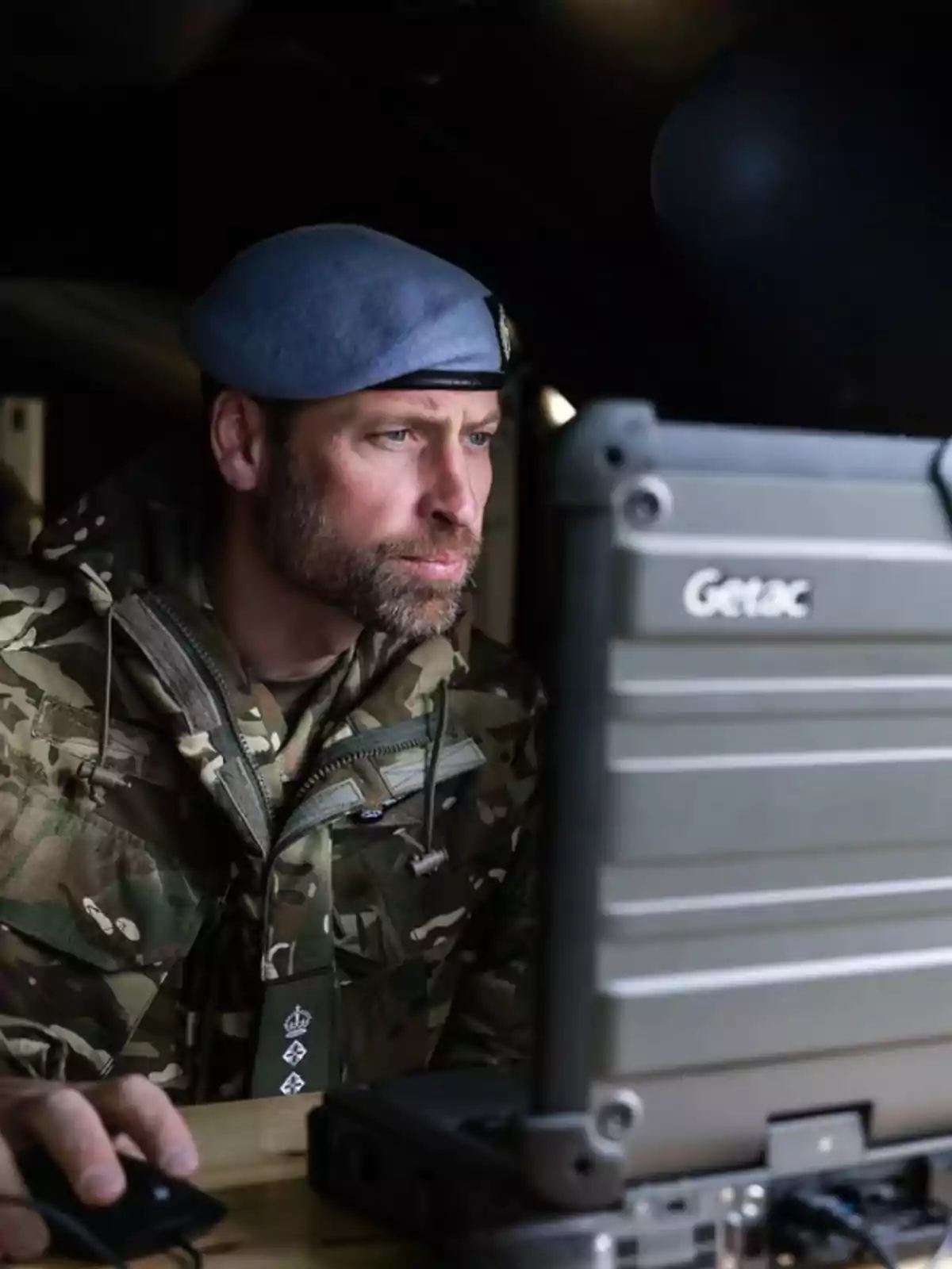 Hombre con uniforme militar y boina azul trabajando concentrado en una computadora portátil robusta Hombre con uniforme militar y boina azul trabajando concentrado en una computadora portátil robusta