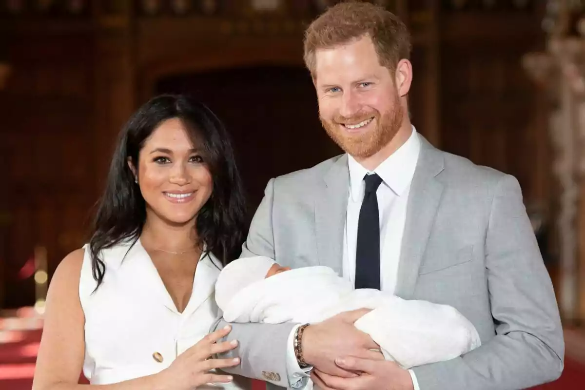 Una pareja, formada por Meghan Markle y el príncipe Harry, sonriente sostiene a un bebé envuelto en una manta blanca.