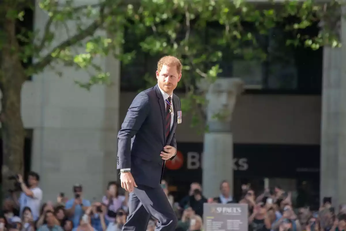 El príncipe Harry con traje oscuro camina al aire libre mientras es fotografiado por una multitud. El príncipe Harry con traje oscuro camina al aire libre mientras es fotografiado por una multitud.