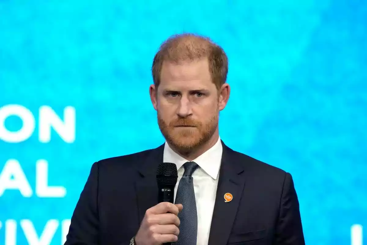 Prince Harry with a beard and dark suit holding a microphone in front of a blue background. Prince Harry with a beard and dark suit holding a microphone in front of a blue background.