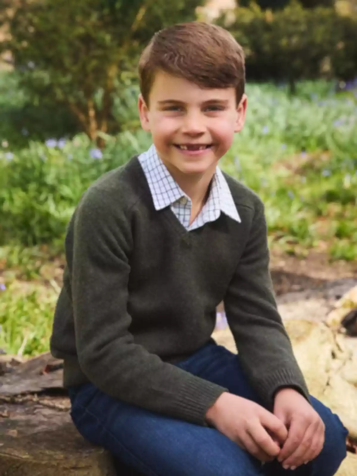 Niño sonriente sentado al aire libre con suéter verde y camisa de cuadros en un entorno natural Niño sonriente sentado al aire libre con suéter verde y camisa de cuadros en un entorno natural