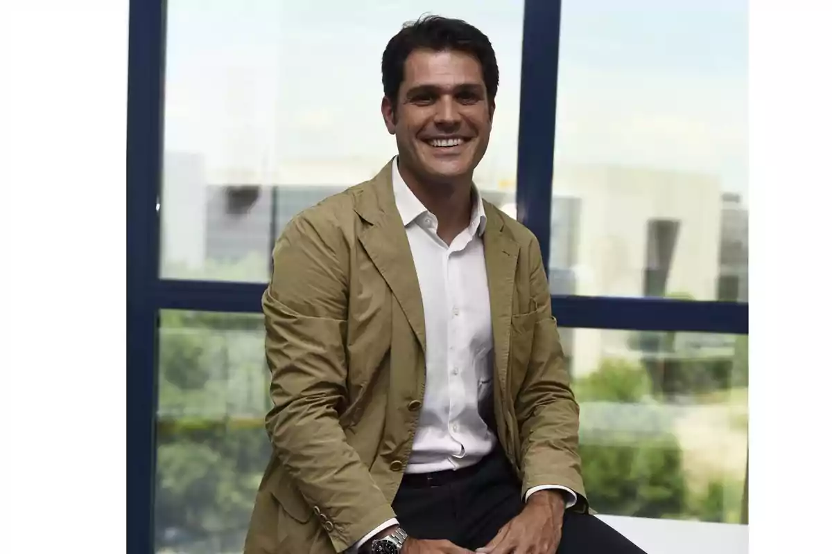 Hombre sonriente con saco beige y camisa blanca sentado frente a una ventana grande