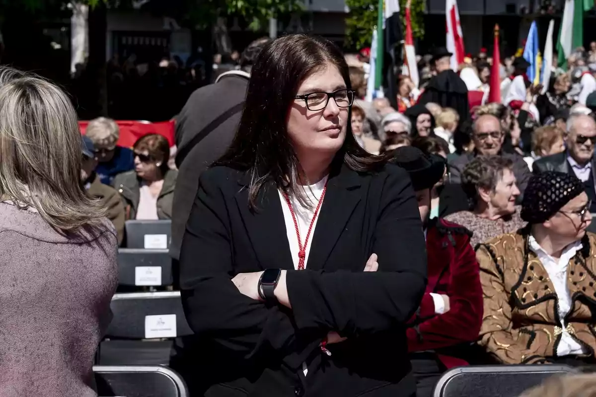 Una mujer con gafas y chaqueta negra está de pie con los brazos cruzados en un evento al aire libre rodeada de personas sentadas y banderas en el fondo. Una mujer con gafas y chaqueta negra está de pie con los brazos cruzados en un evento al aire libre rodeada de personas sentadas y banderas en el fondo.