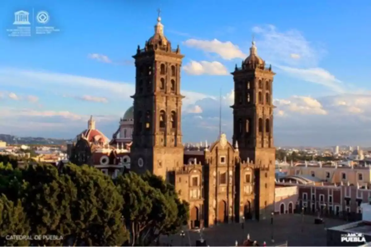 Image of the Puebla Cathedral with its two towers standing out under a blue sky, surrounded by historic buildings and vegetation.