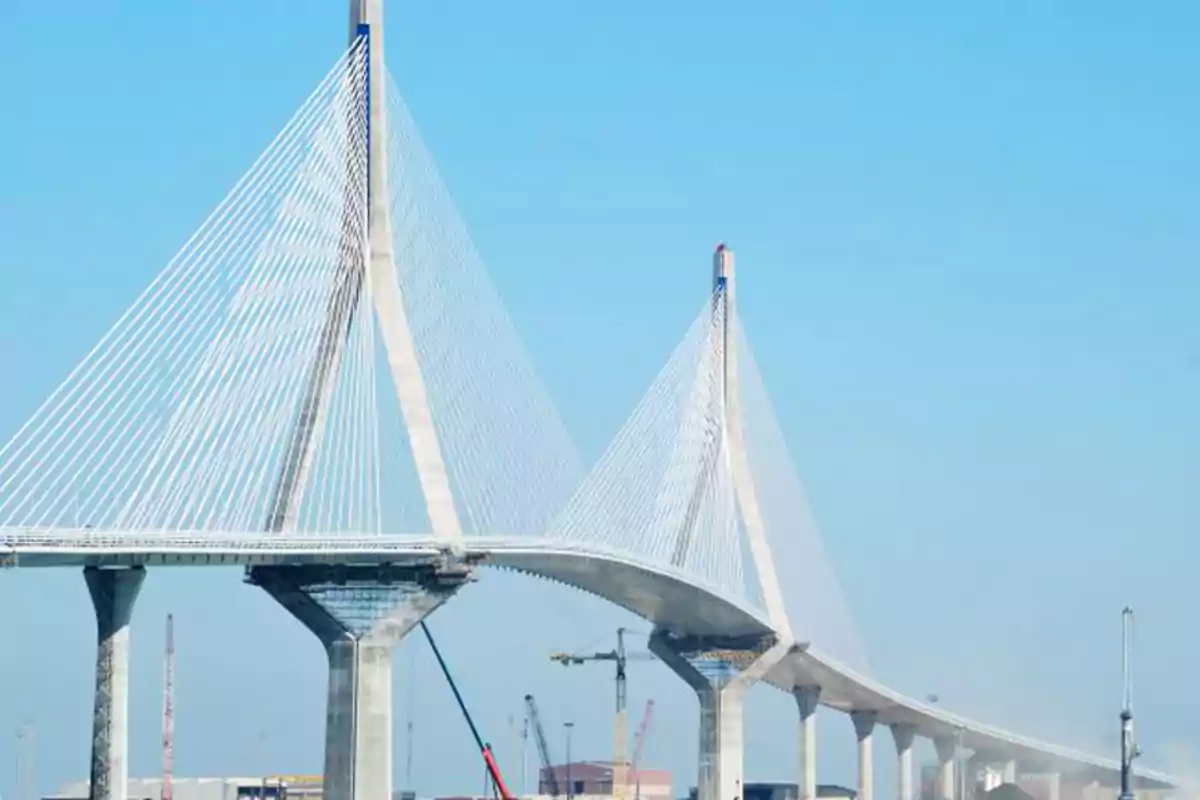 Un puente atirantado sobre un río con grúas y edificios al fondo en un día despejado.